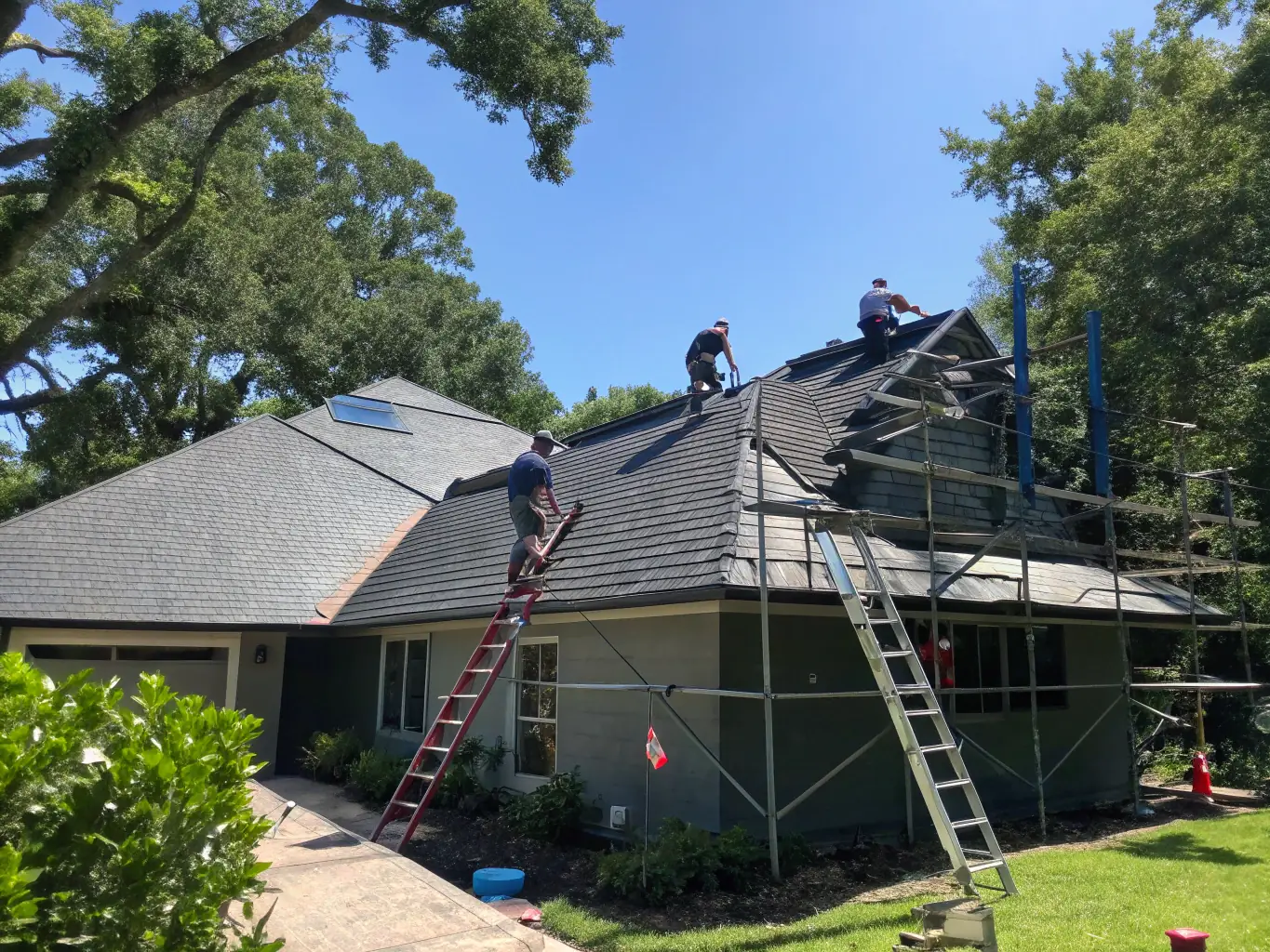 A professional roofing team installing a new shingle roof on a suburban home with clear blue skies in the background, representing TJB Construction's residential roofing services.